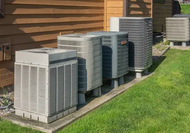 five HVAC units outside against a brown siding and resting on concrete with a green yard around them