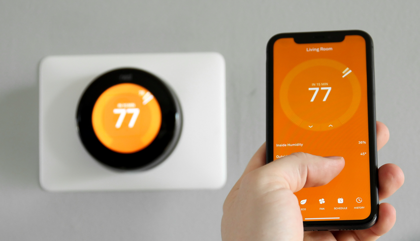 A person using a smartphone app to control a smart thermostat set to 77 degrees in a modern home. Keywords: smart thermostat, energy efficiency, home automation.