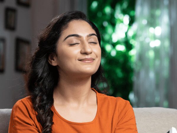 A relaxed woman in an orange shirt sits comfortably with her eyes closed, enjoying the fresh air inside her home. This image represents the importance of Indoor Air Quality for creating a healthy and comfortable living environment.