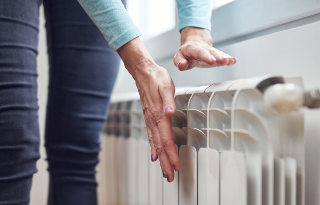 A person holds their hands above a white radiator, feeling the heat, with a window in the background.