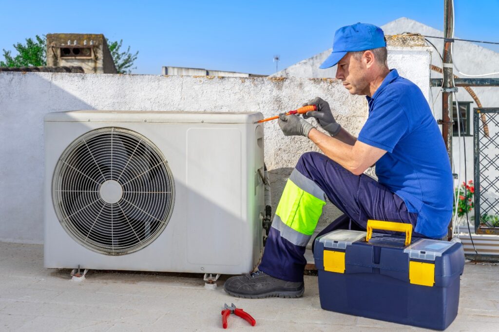 A technician in uniform and gloves uses a screwdriver to repair an outdoor air conditioning unit, with a toolbox and pliers nearby. emergency ac repair