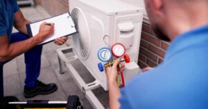 Two technicians service an outdoor air conditioning unit; one checks pressure gauges while the other writes notes on a clipboard. emergency ac repair